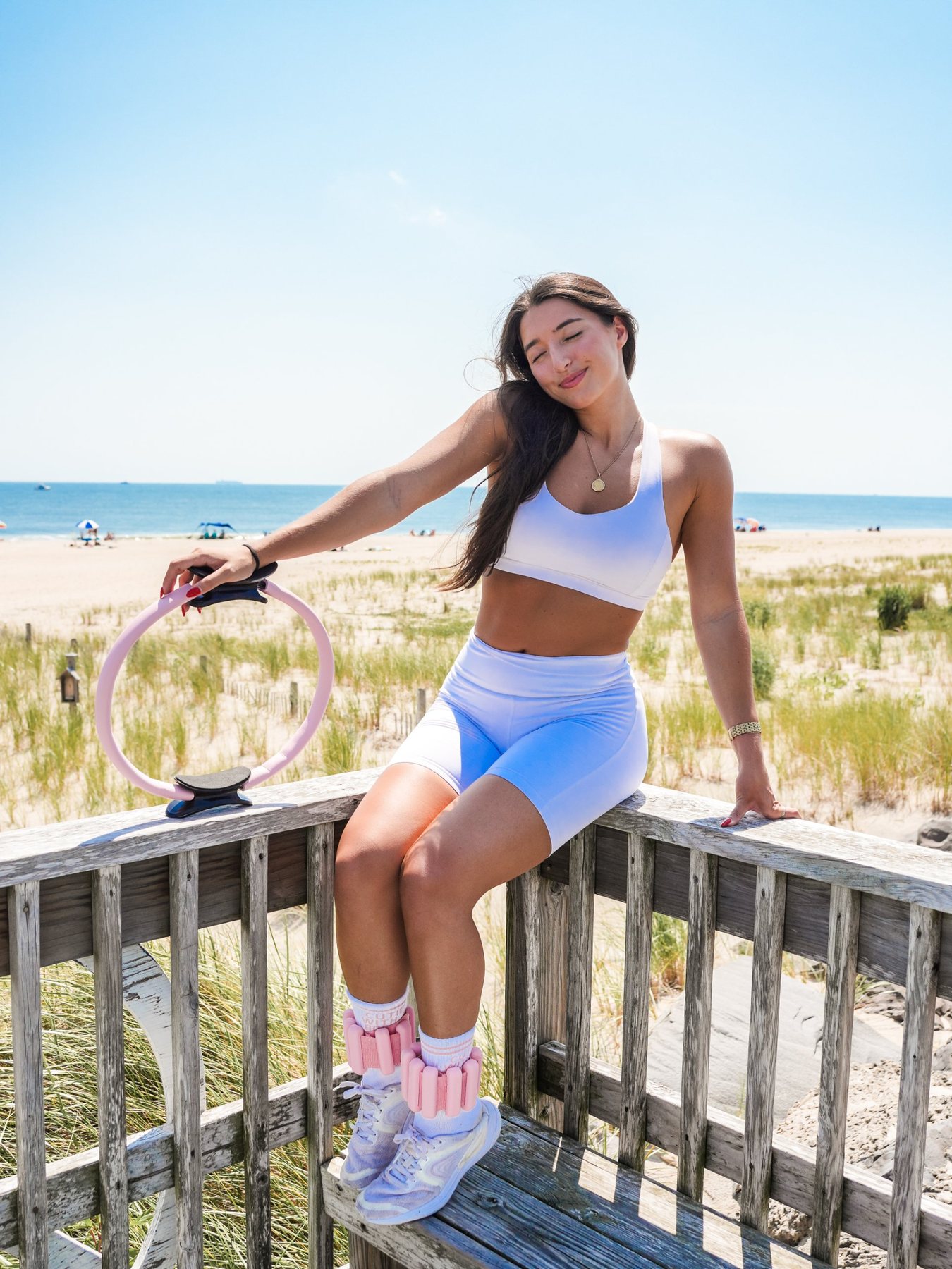 Amanda sitting on a beach boardwalk railing with a Pilates ring and ankle weights